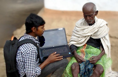 A student uses a laptop to record an interview of an elderly villager for a community project comparing life 25 years ago with the present, documenting tribal folklore and history >A student uses a laptop to record an interview of an elderly villager for a community project comparing life 25 years ago with the present, documenting tribal folklore and history